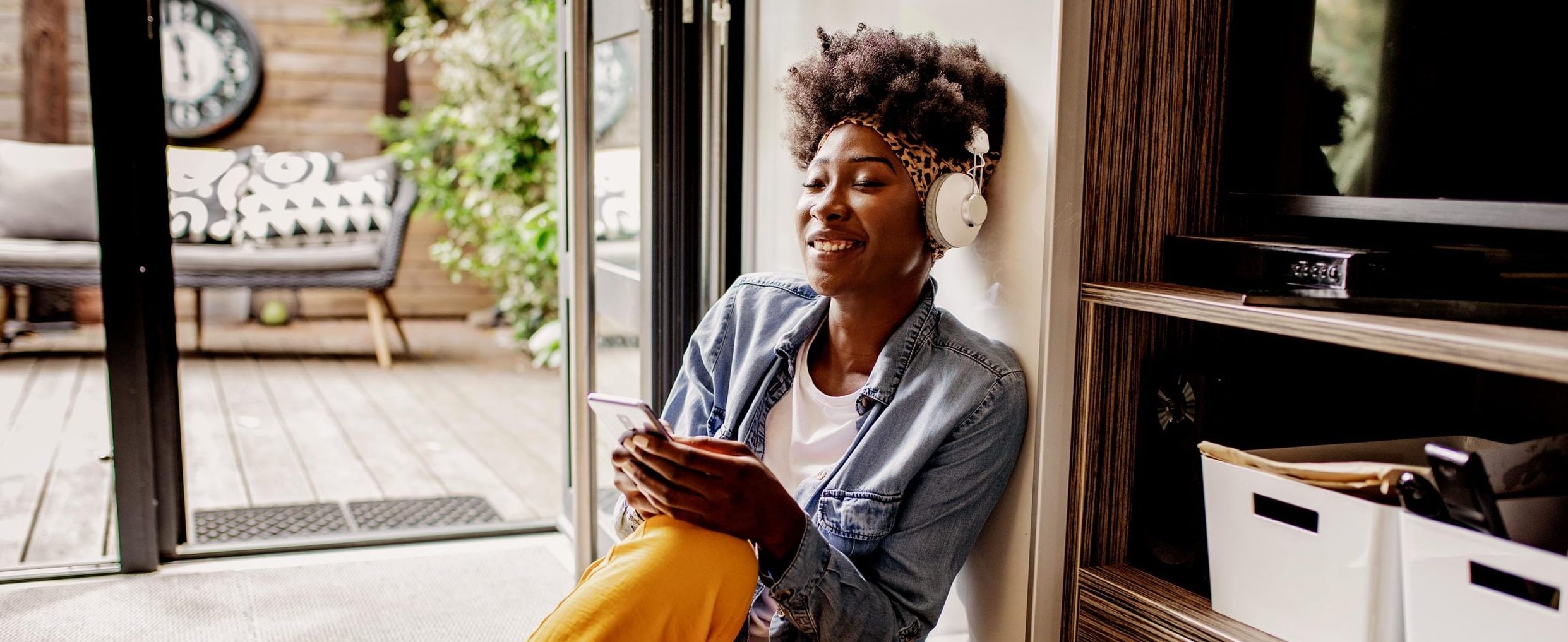 A woman sits by an open door to her home’s patio, wearing headphones and smiling at her mobile phone