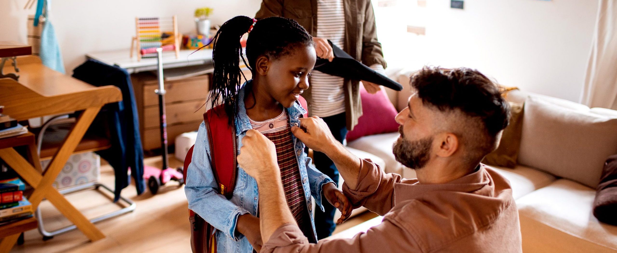 A man smiles while helping his young daughter get ready for school, with a couch and desk in the background