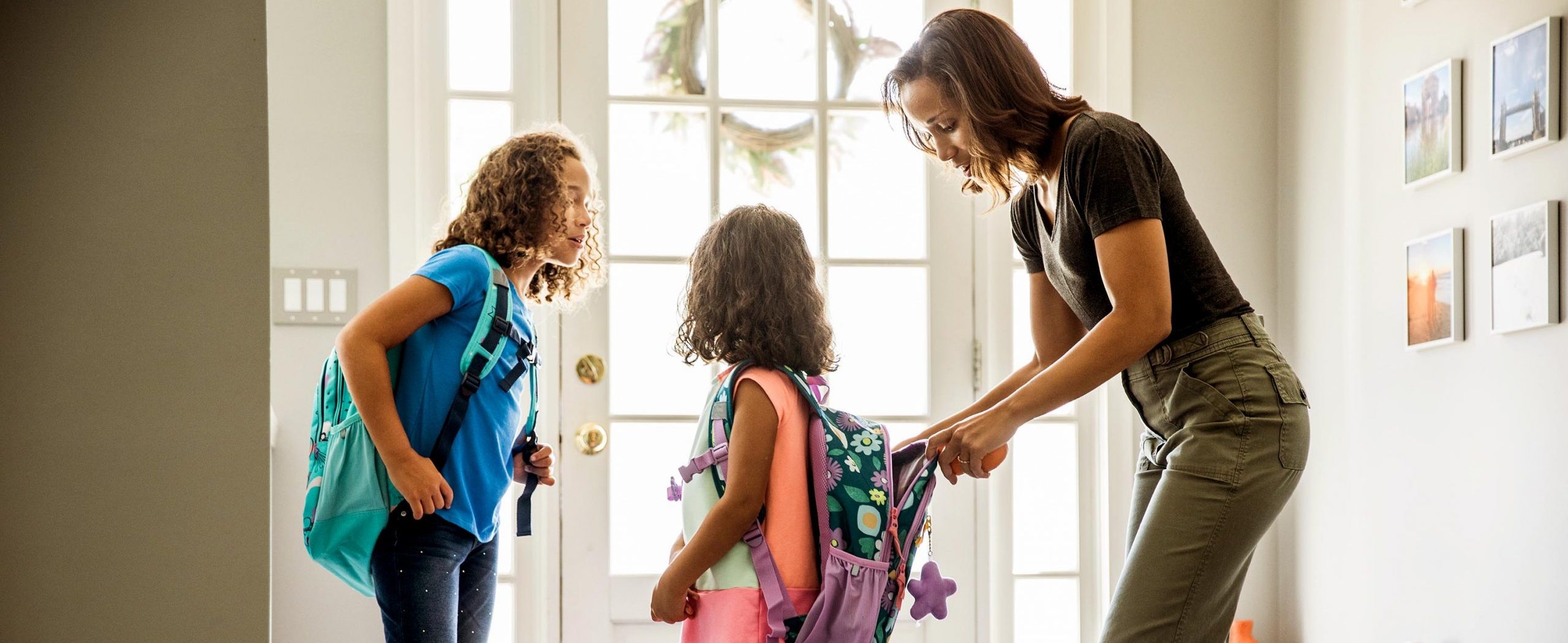 Standing in an entryway, a woman zips her young daughter’s backpack as another daughter looks on.
