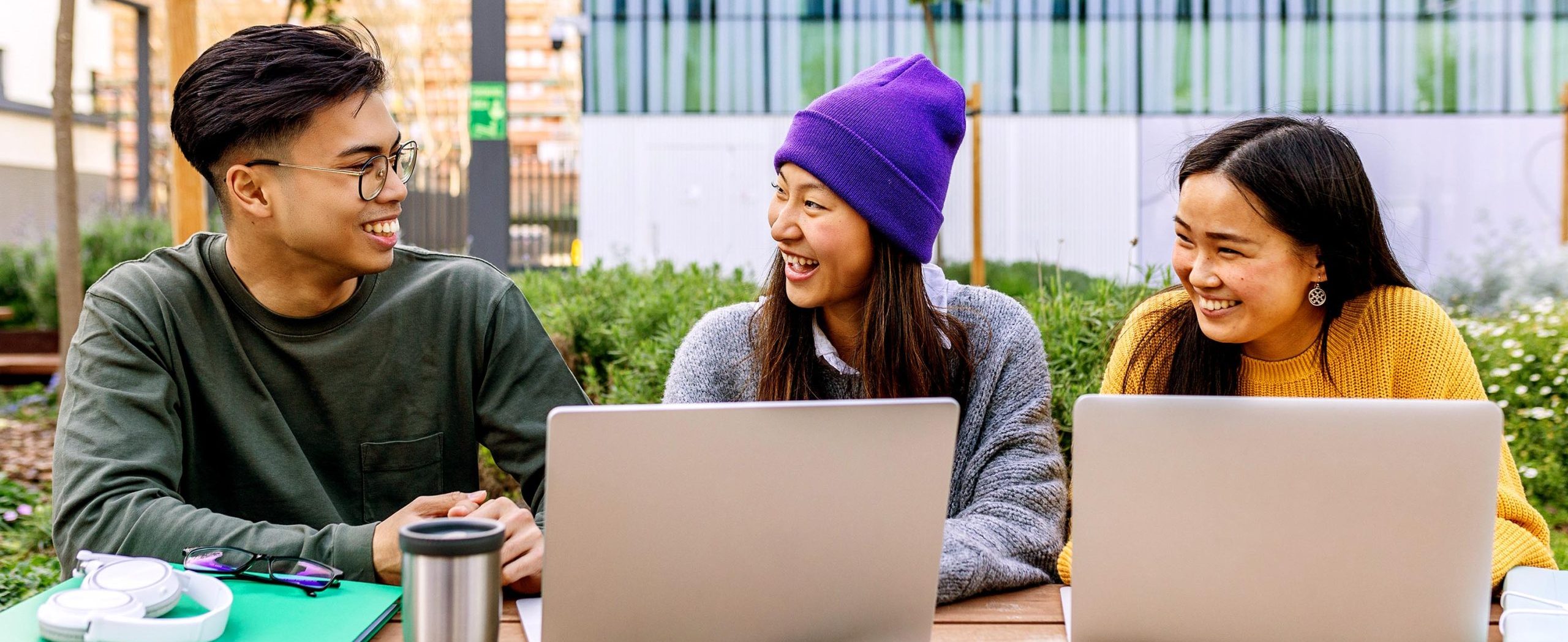 Three college students sit outside and talk, next to laptop computers and coffee.