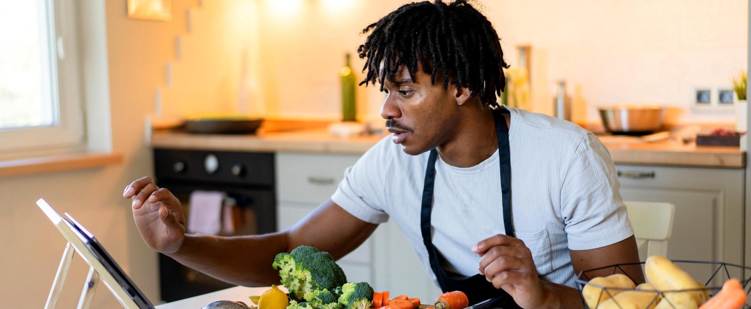 A young man reads a recipe on his tablet device, next to containers with fish, carrots, and broccoli on a kitchen counter.