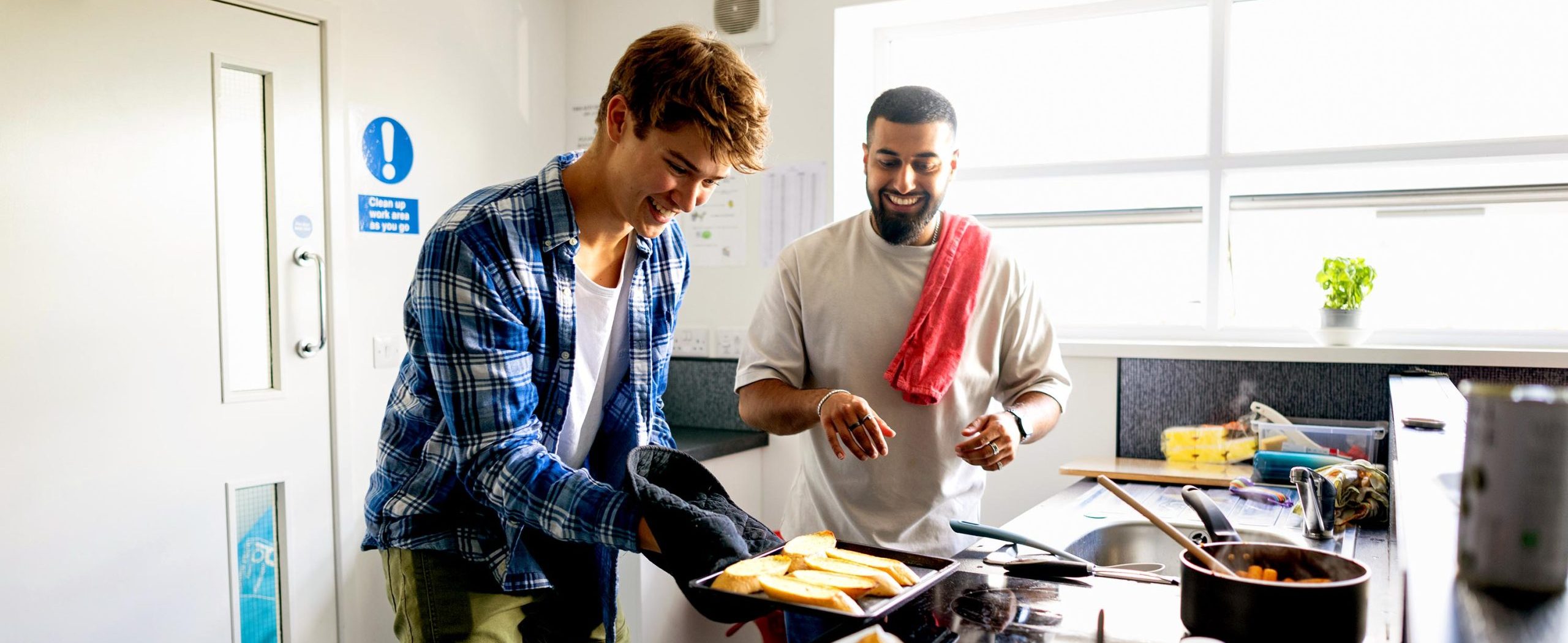 Two young men smile while cooking, one holding a pan of sliced bread with an oven mitt.