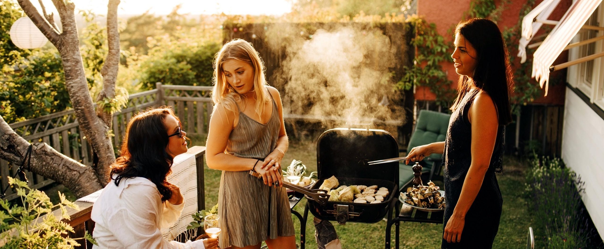 Three women chat while grilling in a sunny backyard.