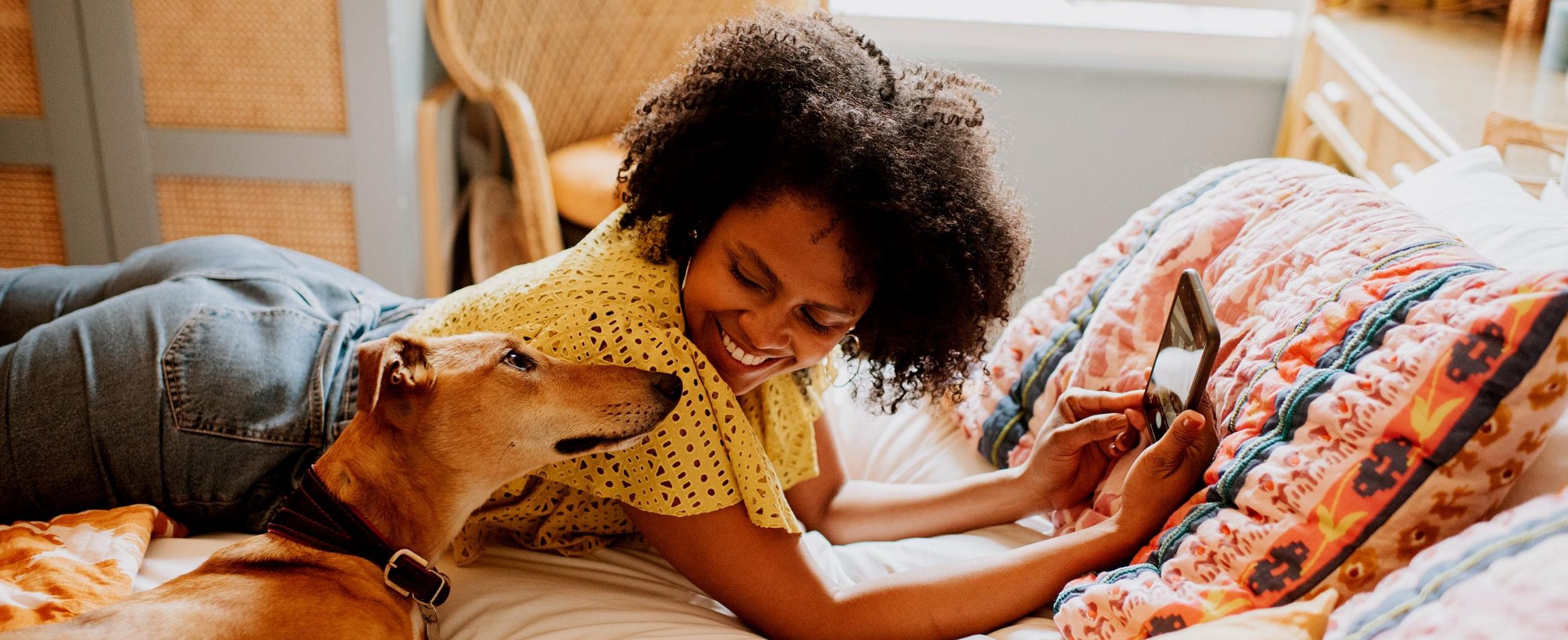 A woman relaxes on a bed with her dog while looking at her mobile phone.