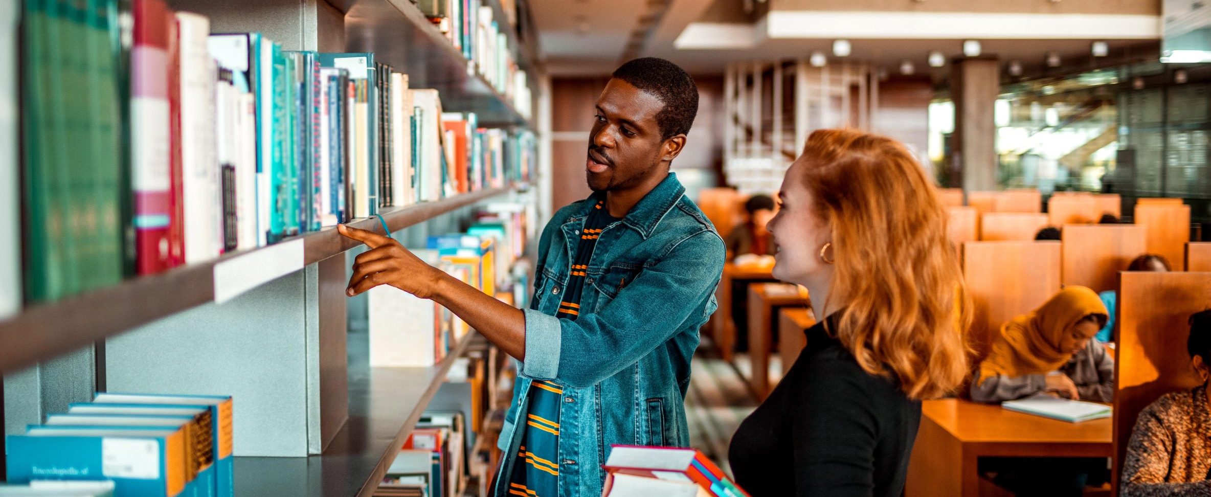 Two college students search for books in the library, while other students study in the background.