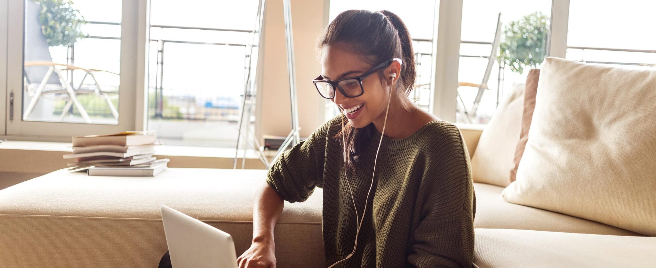 A young woman wearing headphones smiles at her laptop computer, with a pile of books on a couch in the background.