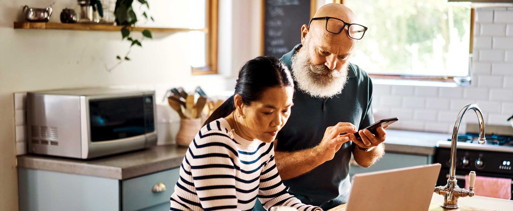 A woman sits at a kitchen counter with her laptop, next to a man holding a mobile phone.