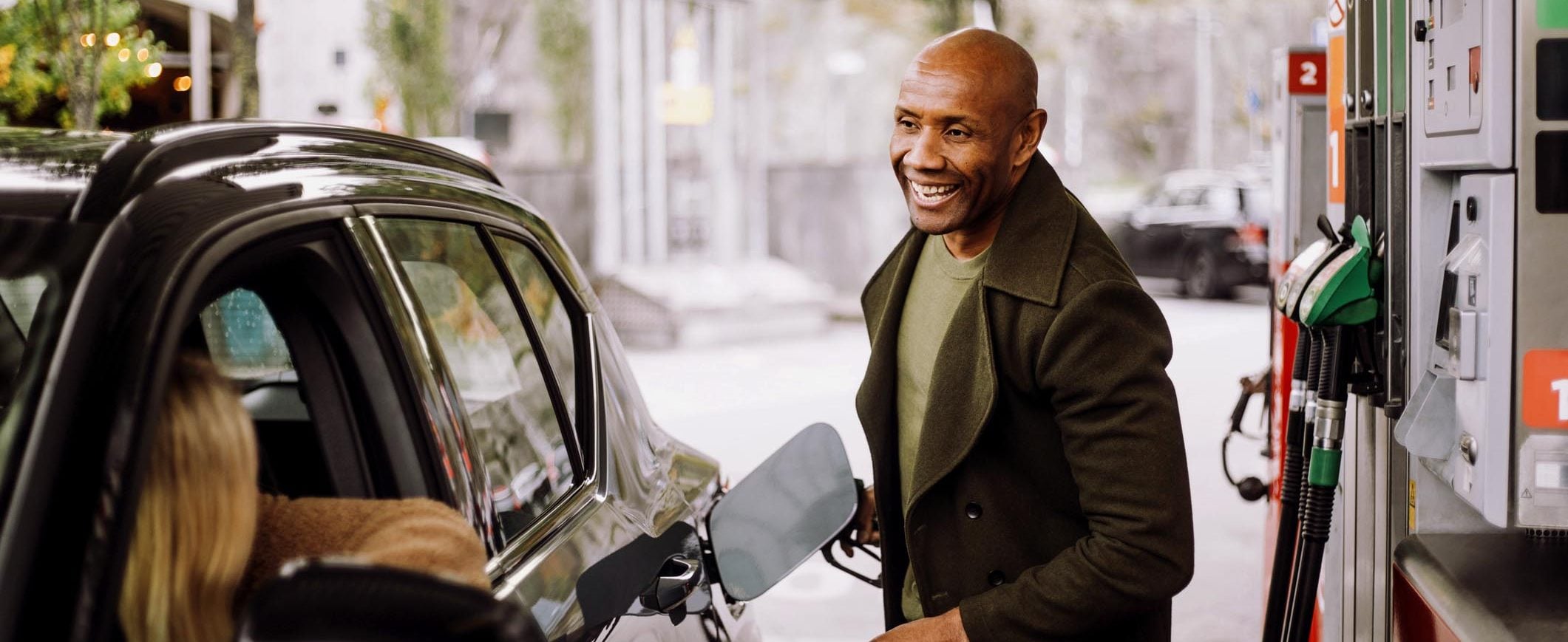 At a gas pump, a man fills the car’s tank while talking with a woman in the driver’s seat.