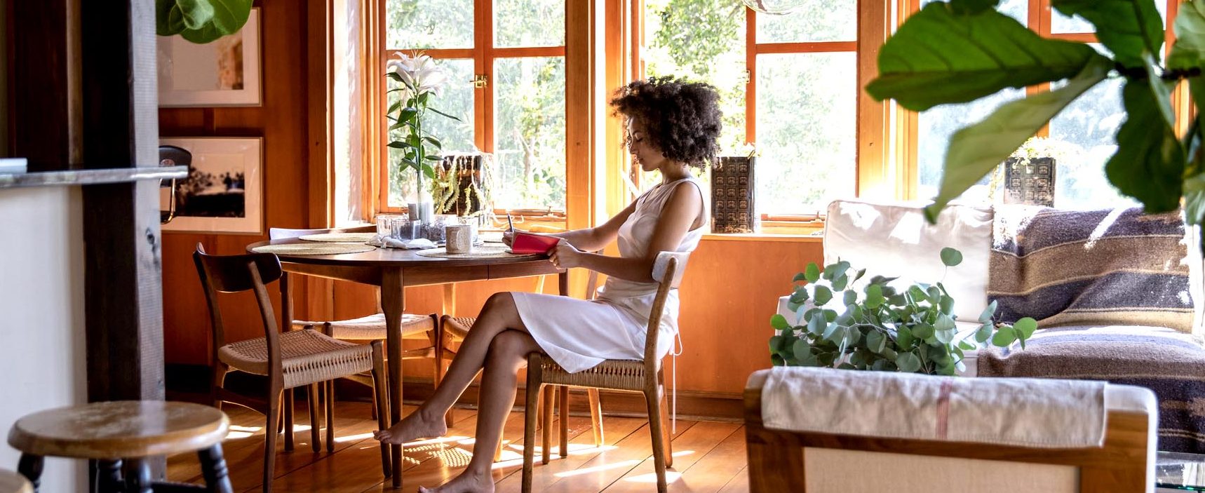 A woman sits at a table and writes in a notebook, with windows and plants in the background.