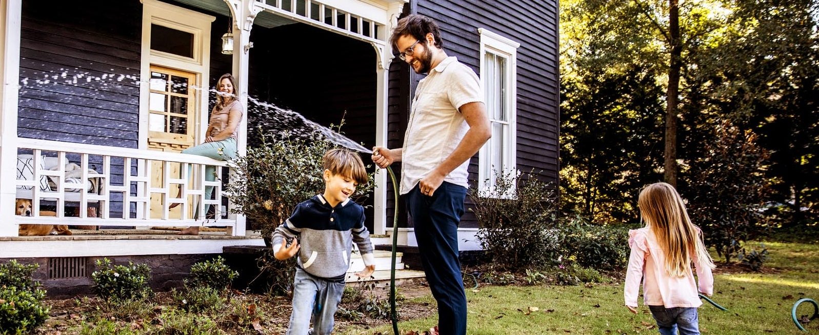 A man smiles and sprays a garden hose while two young children play in the yard.