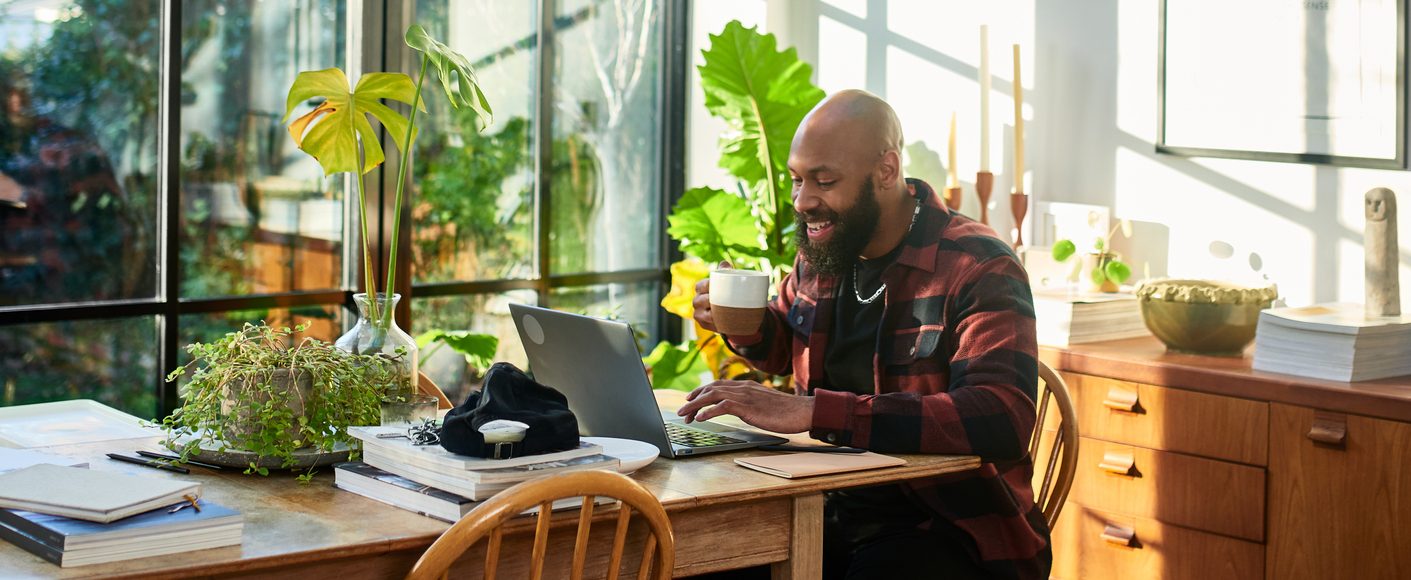 A man holds a mug and works on a laptop computer, with windows and plants in the background.