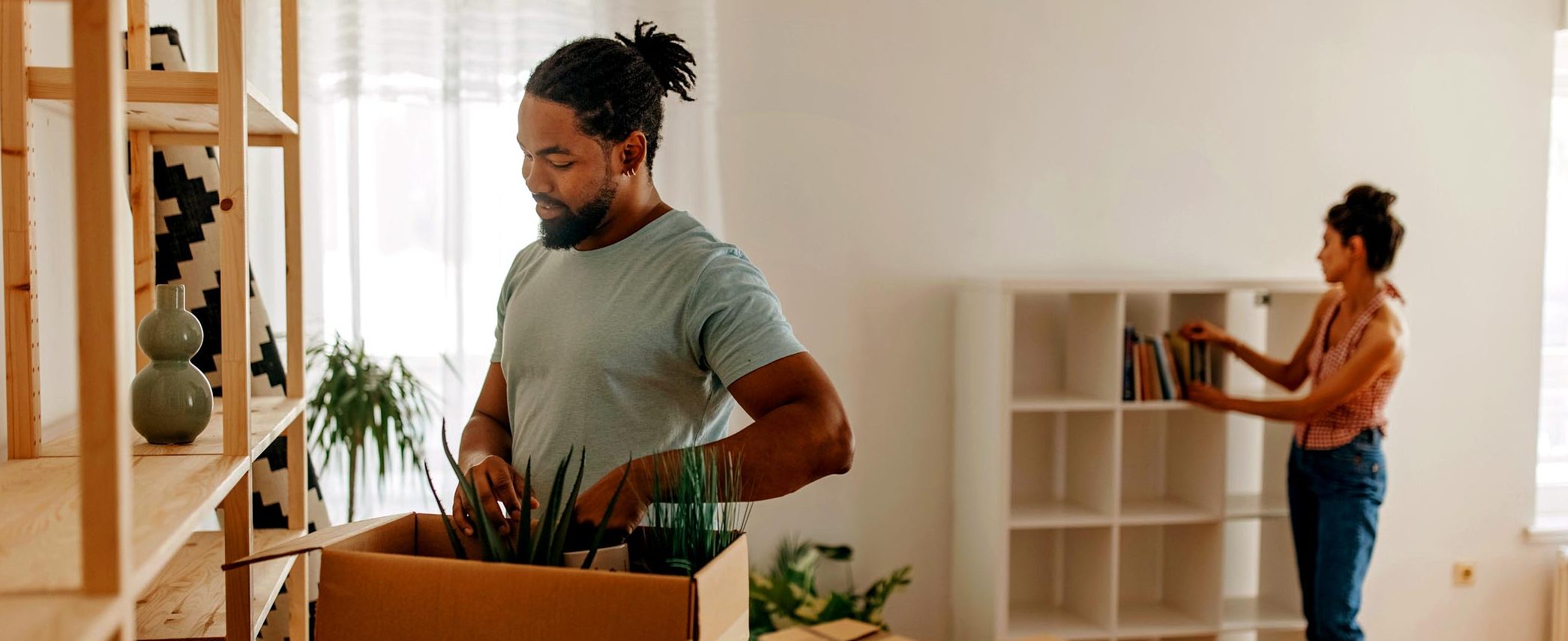 A man stands next to a box filled with plants, while a woman adds books to an empty bookshelf in the background.