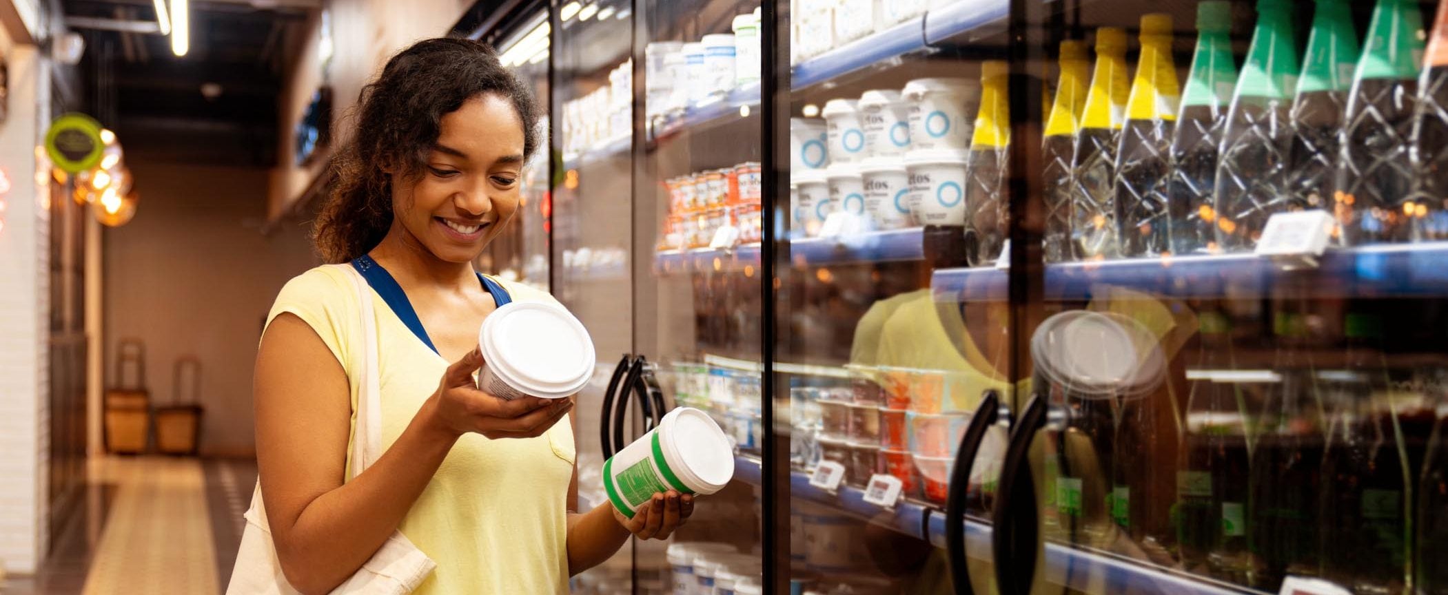 Standing in a grocery aisle, a woman compares two small containers. 