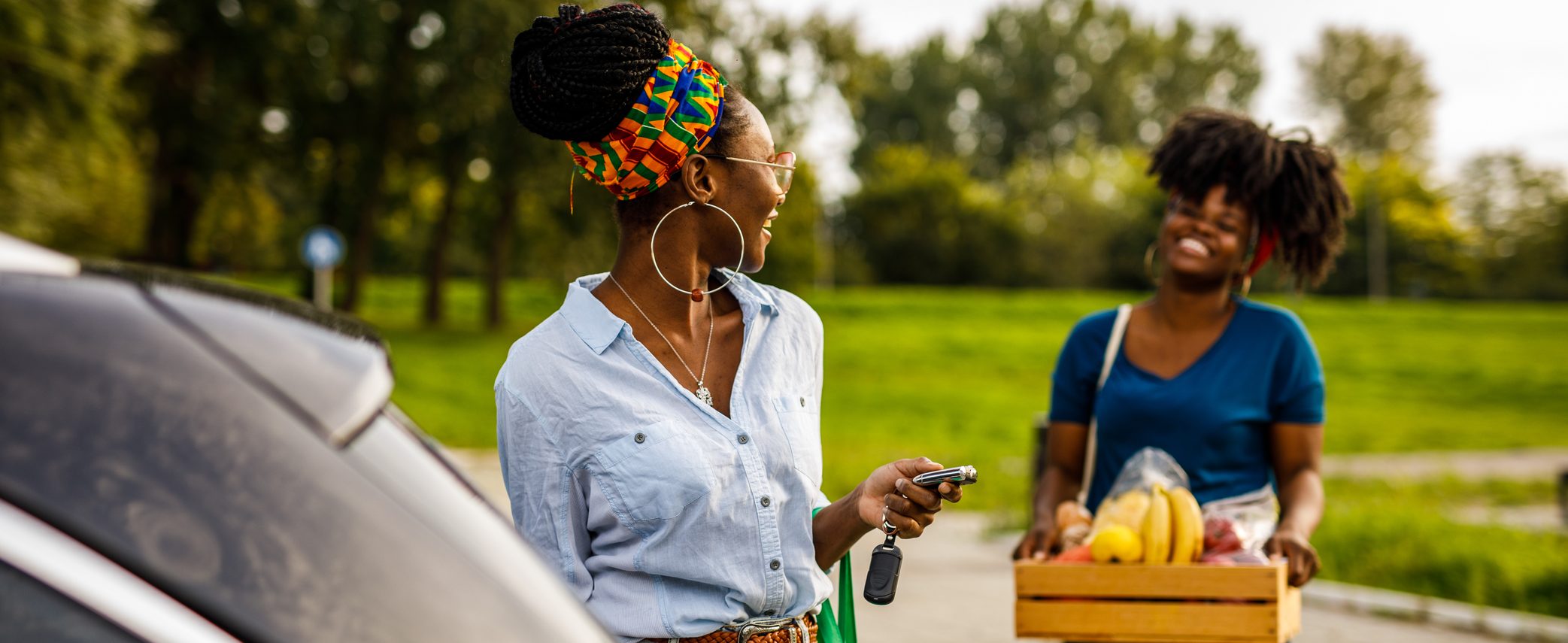 Two women stand next to a car, one carrying a basket of fruits and vegetables.