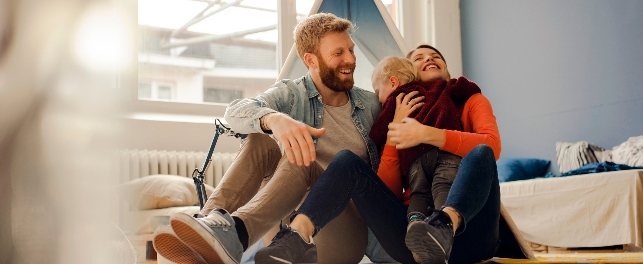 A couple smiles and embraces their young child, with a couch and window in the background.