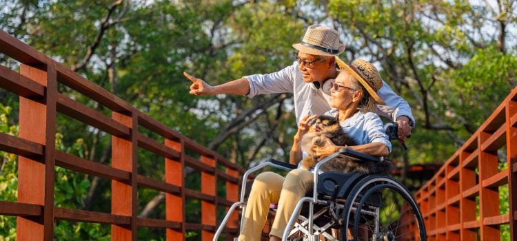 On a bridge, a man stands next to a woman, who’s seated in a wheelchair with a dog in her lap.