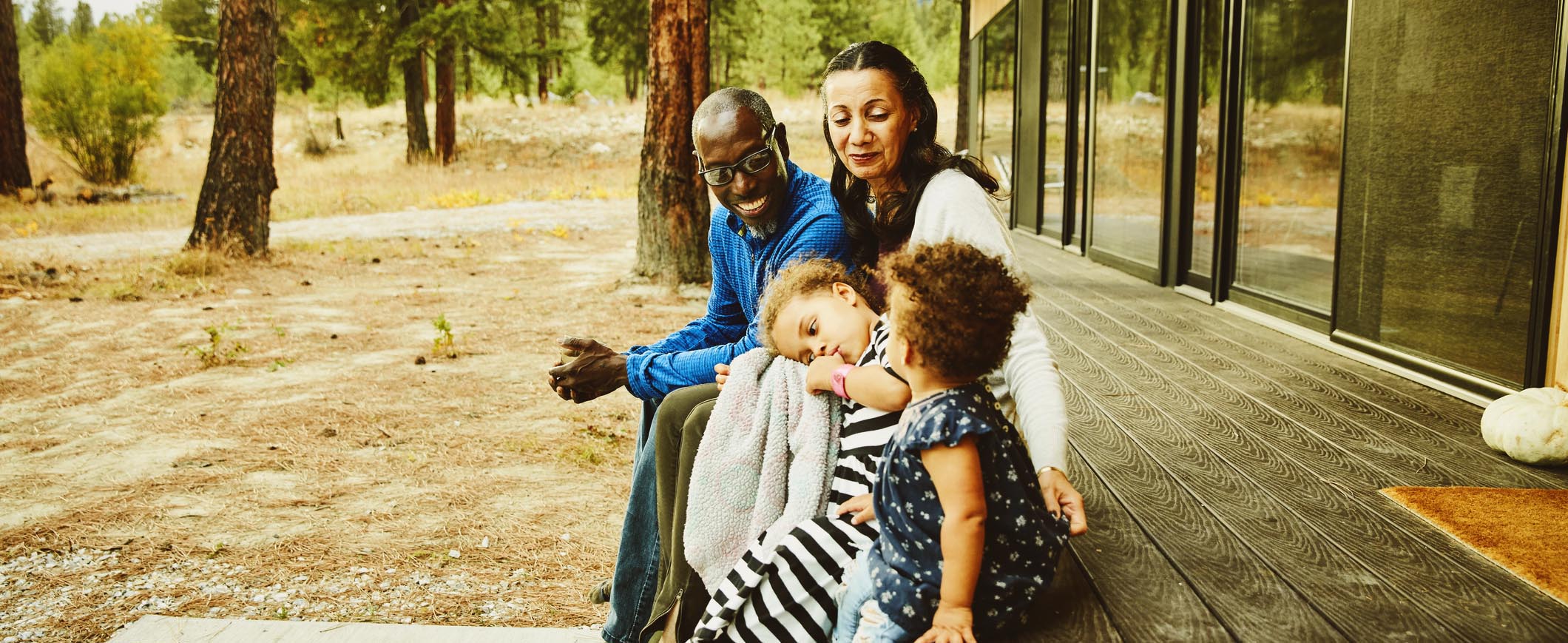 A couple smiles while sitting with their two young children on a patio, with trees in the background.