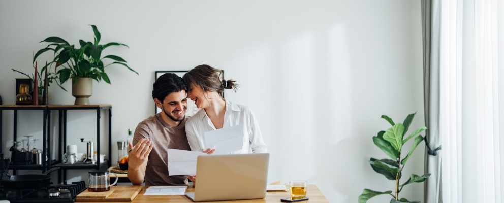 A couple stands close together while they review documents at the kitchen table.