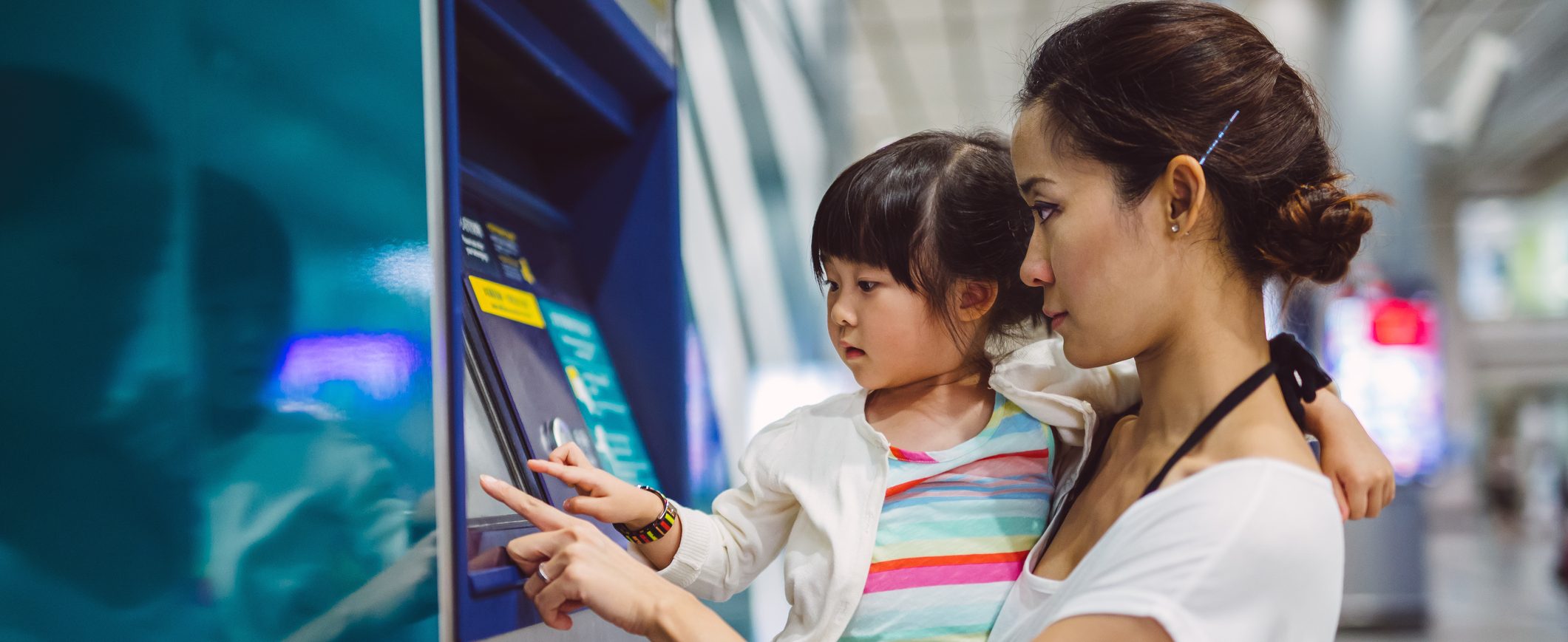 A woman holds her young daughter as they both press an ATM screen.