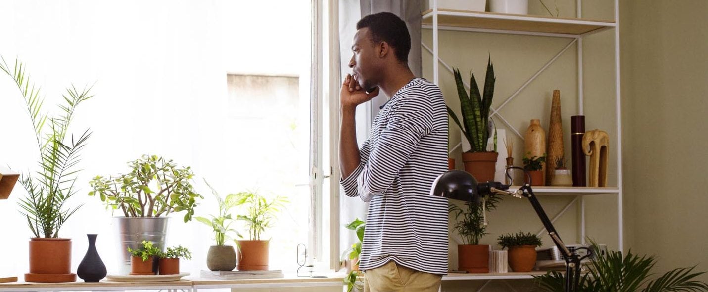 A man stands and looks out a window in deep thought, next to bookshelves and plants.