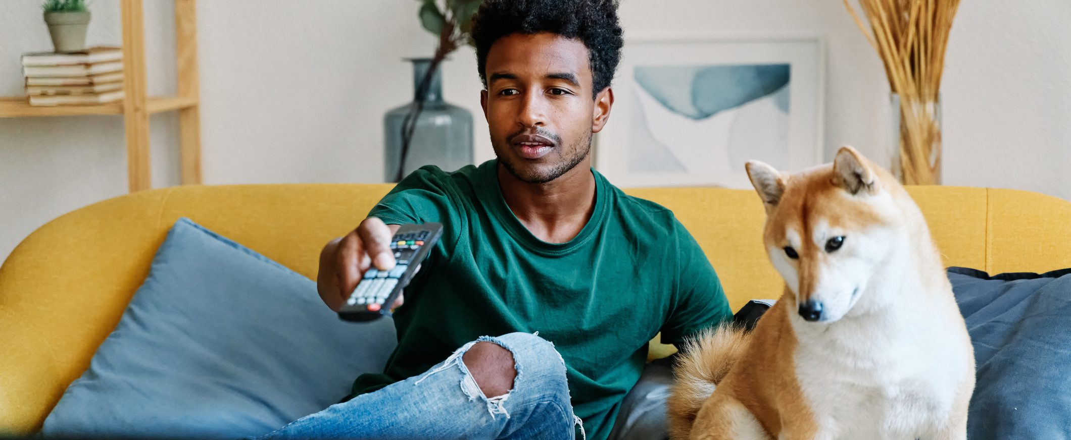 A man sits on a couch with his dog, pointing a remote control.