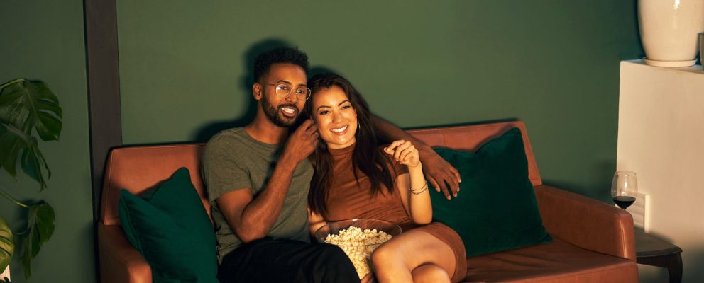 A couple sits on a couch while watching a movie and eating popcorn.