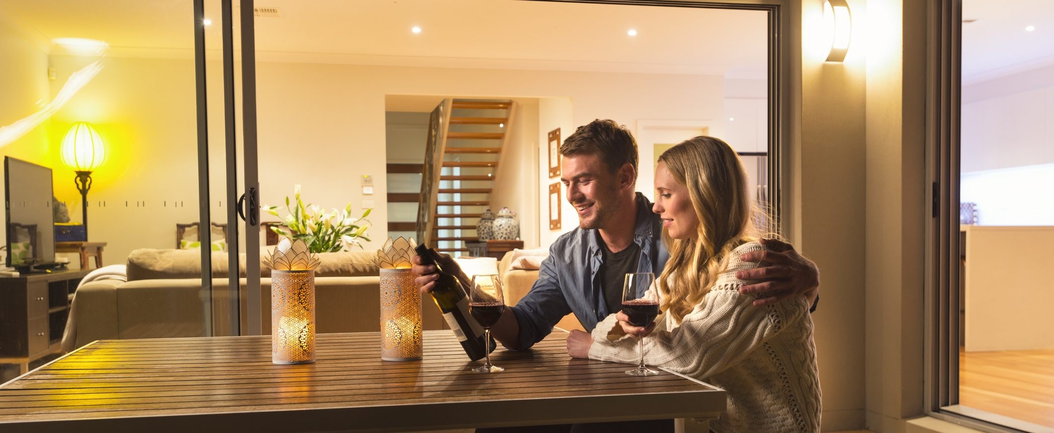 A couple sits at a wooden table on a porch in the evening, looking at a bottle of wine together.