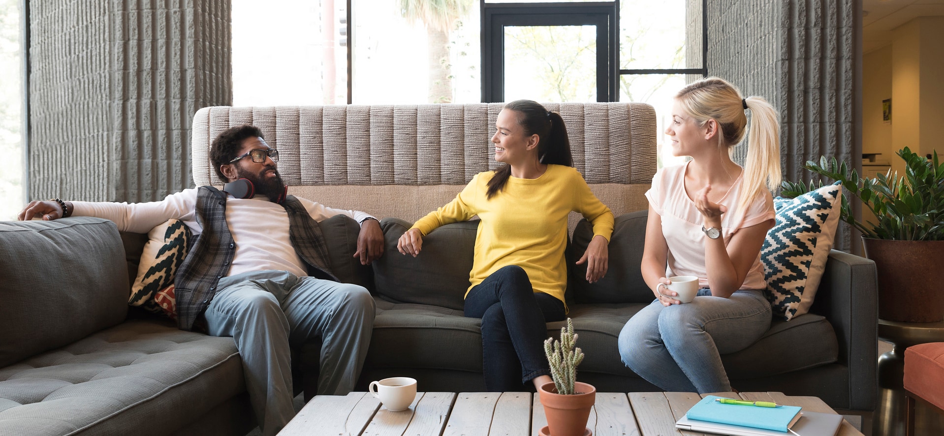 Three friends sit together on a sofa and chat, one holding a cup of coffee.