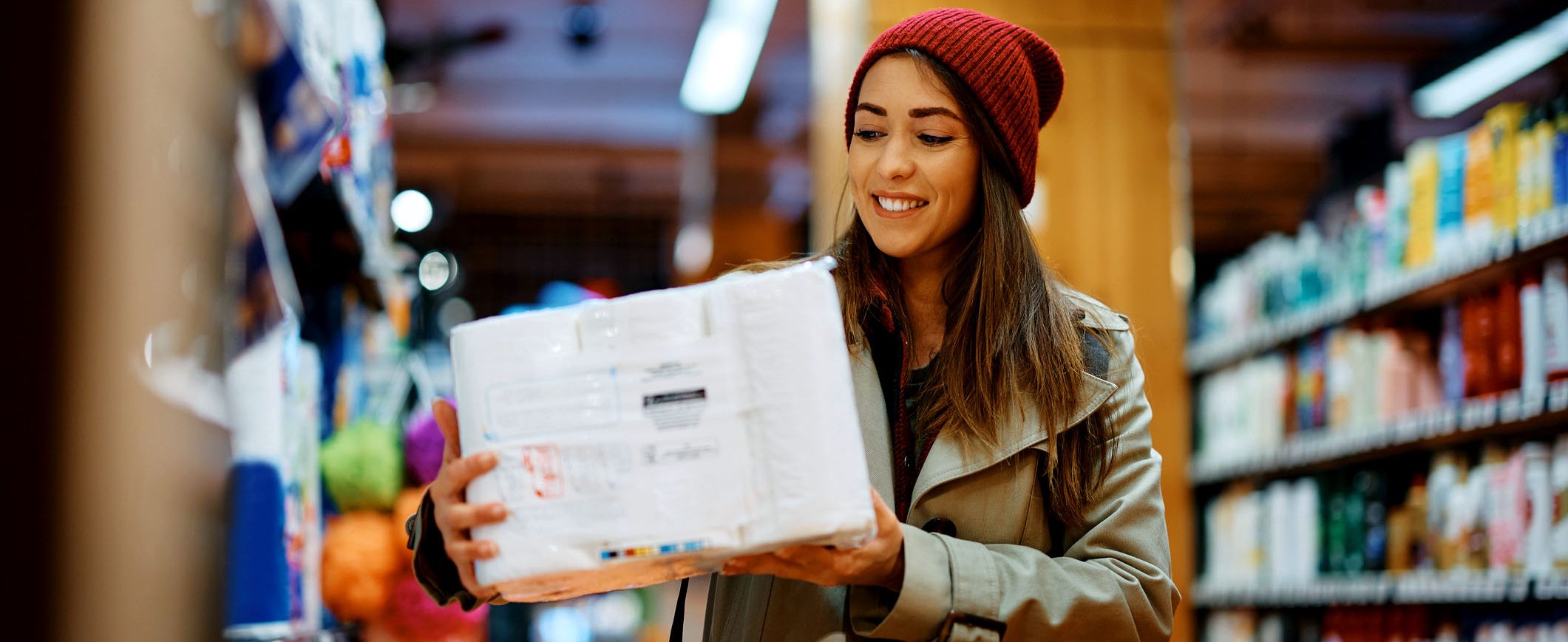 A smiling young woman, wearing a trench coat and winter hat, holds a package of paper towels at the grocery store.