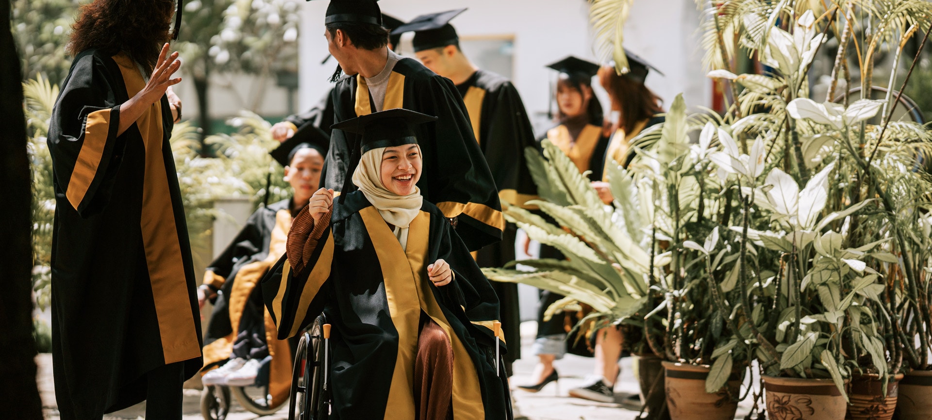Graduates in black and gold robes celebrate outdoors. One woman using a wheelchair and wearing a hijab smiles brightly in the foreground.