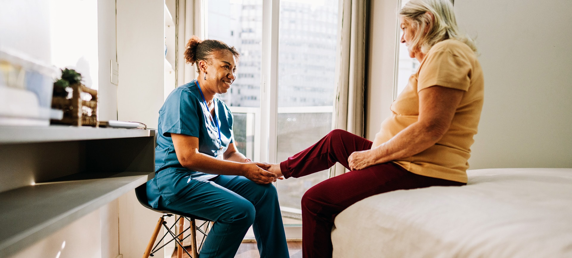 A nurse wearing blue scrubs assists an elderly woman sitting on a bed in a bright, sunny room. Both women smile. 
