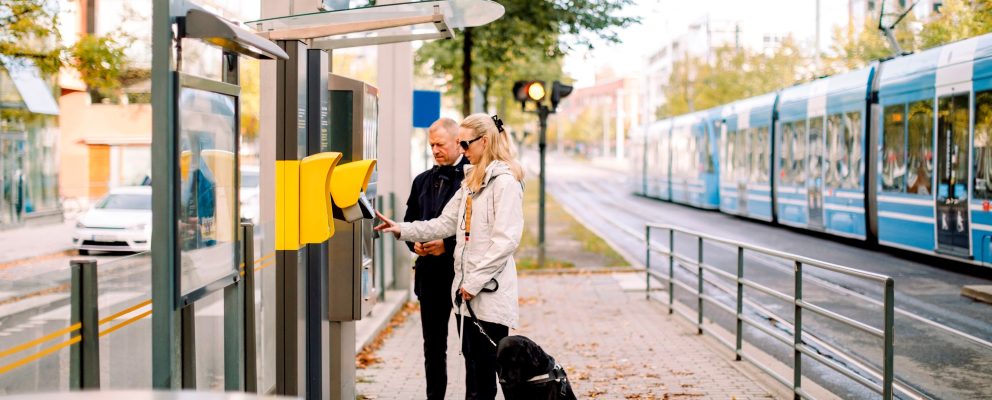 A blind woman uses a keypad outside of a train station while a man stands by her side.
