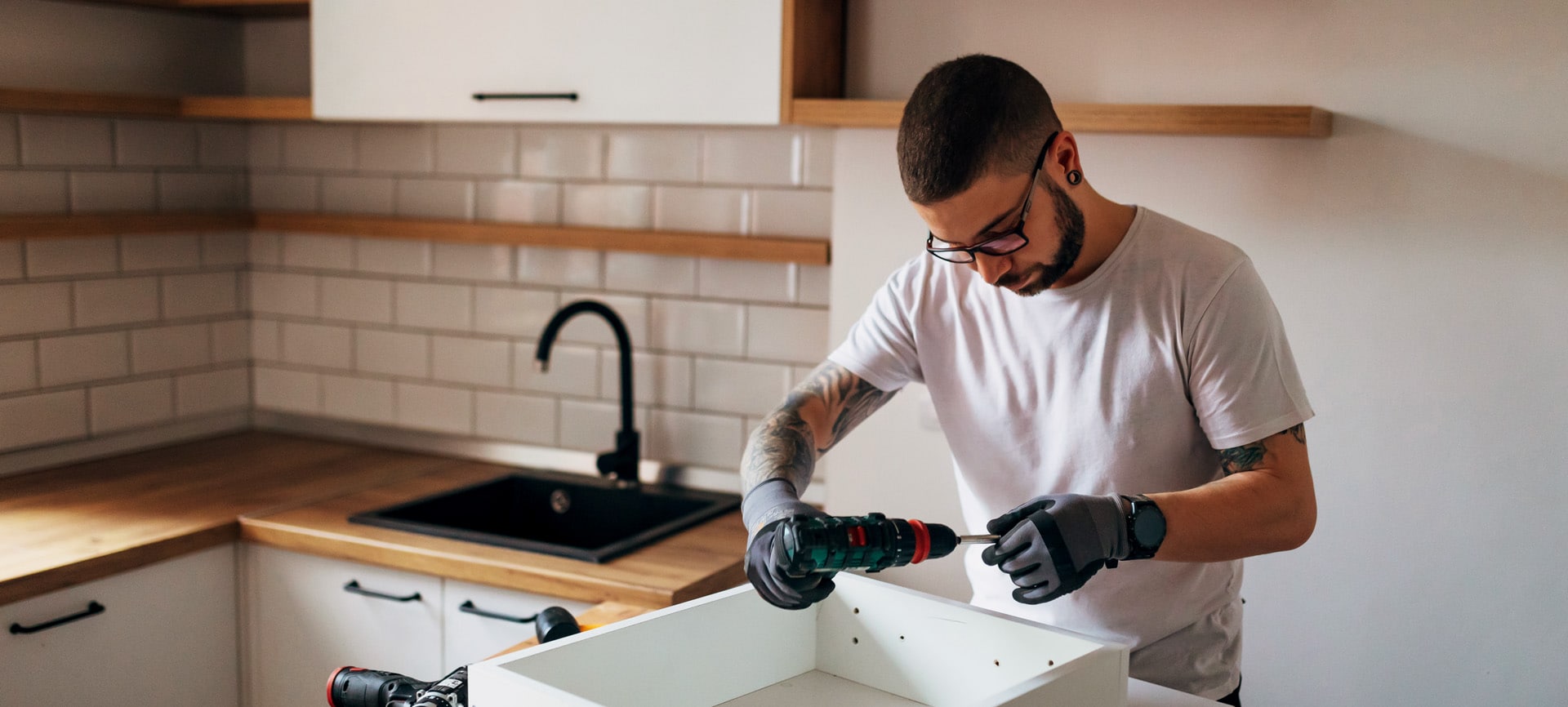 A young man holds a power drill and prepares to assemble a drawer in his kitchen.