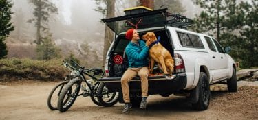 A man sits with his dog in an open car trunk, getting ready to hike a forest trail.