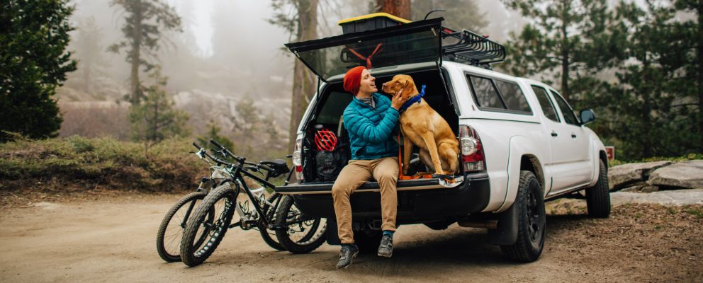 A man sits with his dog in an open car trunk, getting ready to hike a forest trail.