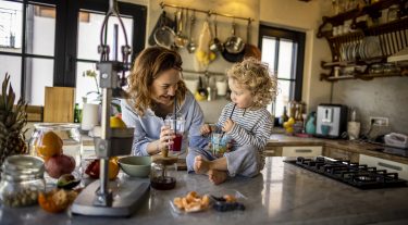 A woman stands in her kitchen holding a drink with straw, next to her young daughter who sits on the counter.