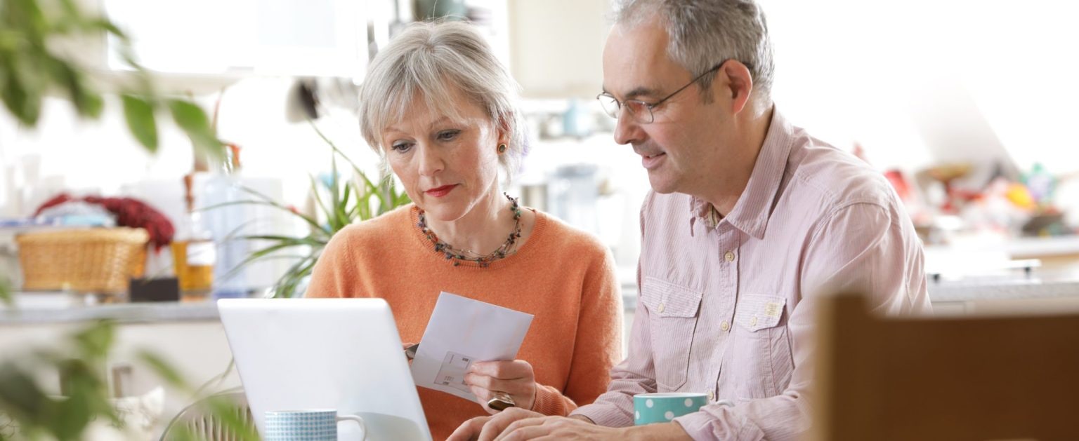 An older couple sit together at a kitchen table and look at a laptop screen together.