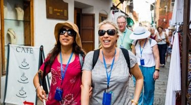 A woman wearing sunglasses and holding a cane walks arm-in-arm with a friend on a city street, accompanied by their tourist group.