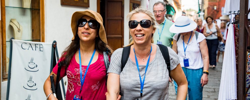 A woman wearing sunglasses and holding a cane walks arm-in-arm with a friend on a city street, accompanied by their tourist group.