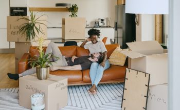 A couple relaxes on a couch surrounded by unpacked moving boxes in their living room.