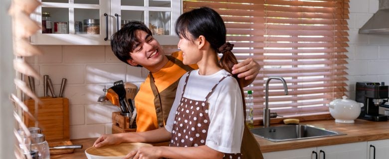 A young couple smiles and looks at each other while cooking in a brightly lit kitchen. 