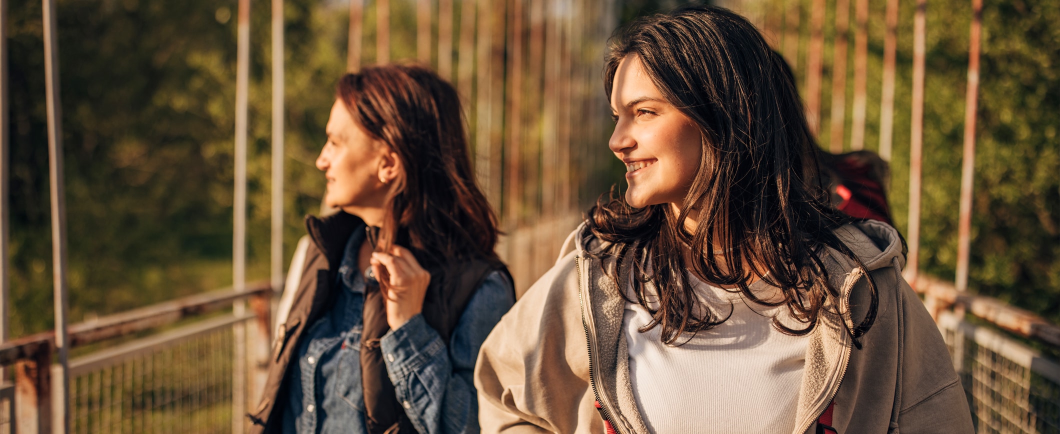 A mother and daughter walk across a bridge while hiking and look into the sun. 