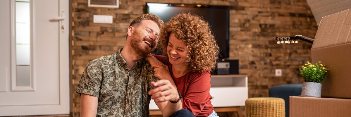 A young couple sits on the floor of their new home, laughing, next to moving boxes.