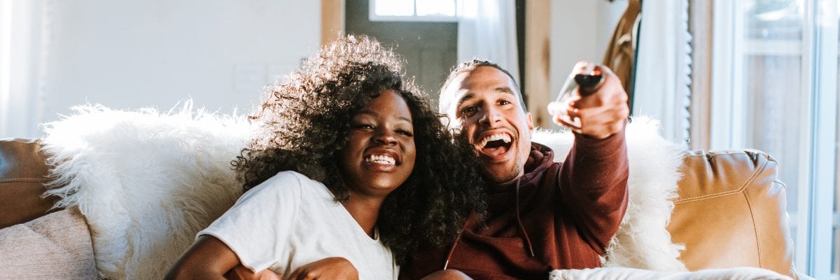 A couple sits together on the couch, laughing while watching TV.