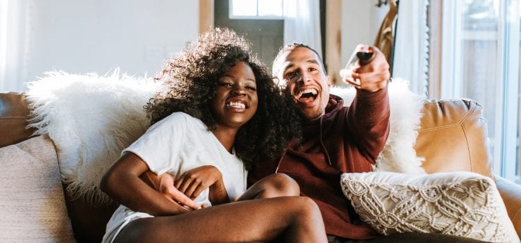 A couple sits together on the couch, laughing while watching TV.