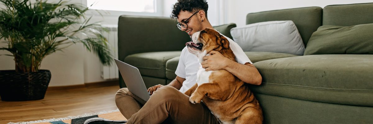 A young man sits on the floor with his laptop, hugging his dog and smiling.