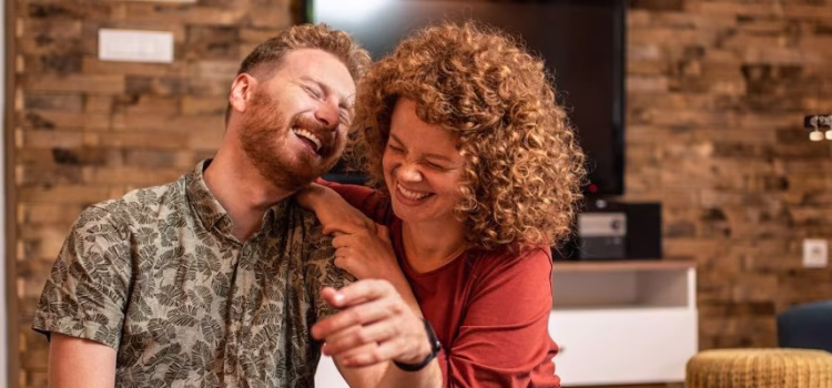 A young couple sits on the floor of their new home, laughing, next to moving boxes.