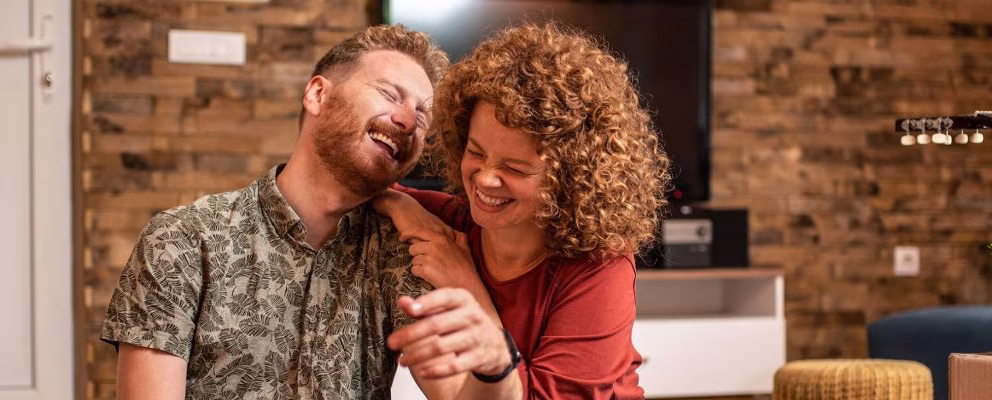 A young couple sits on the floor of their new home, laughing, next to moving boxes.