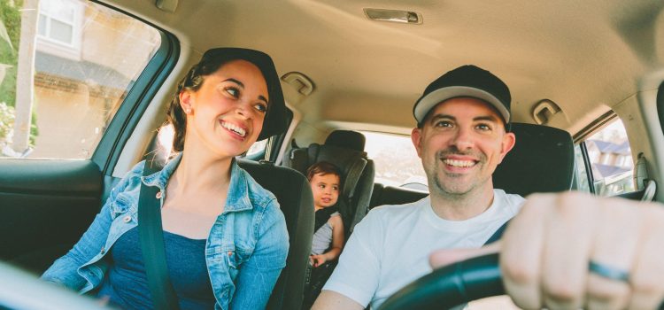 A couple smiles from the front seat of their car as their baby sits in a cars eat in the back.