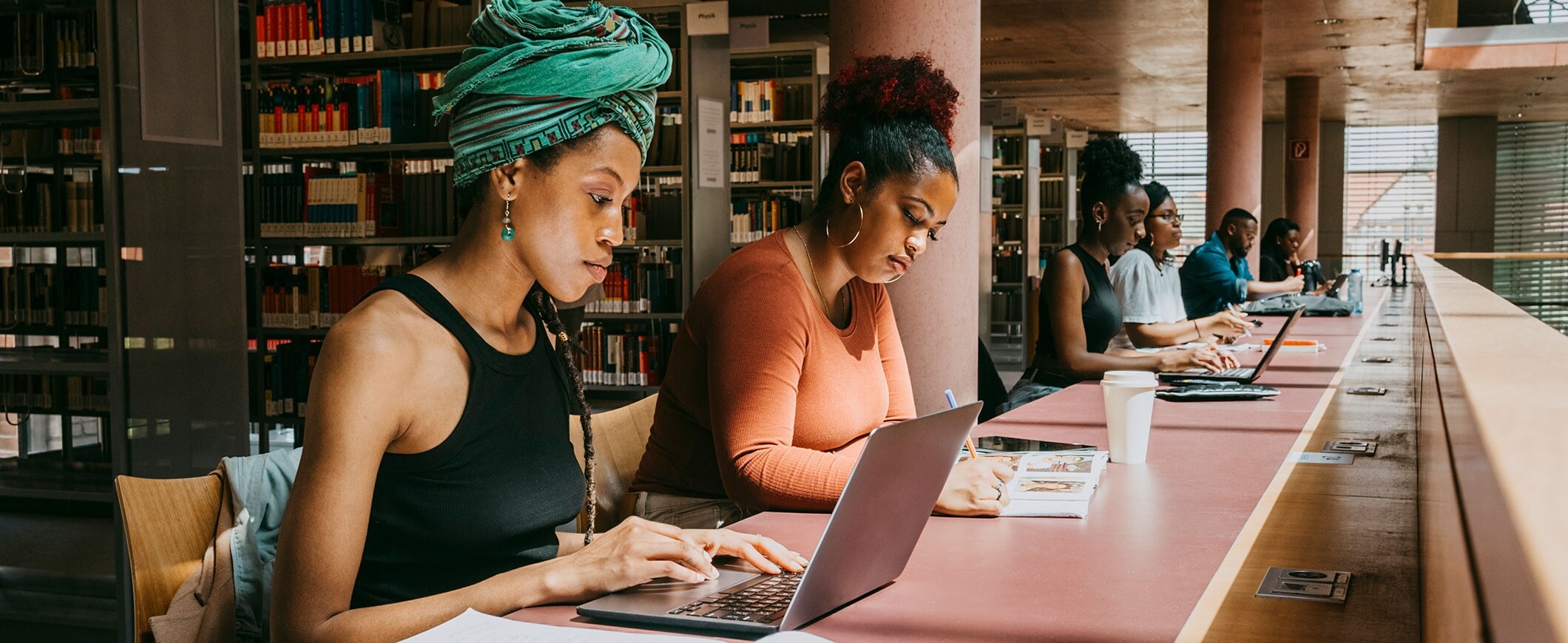Two young women work on laptops at a long table in a library.